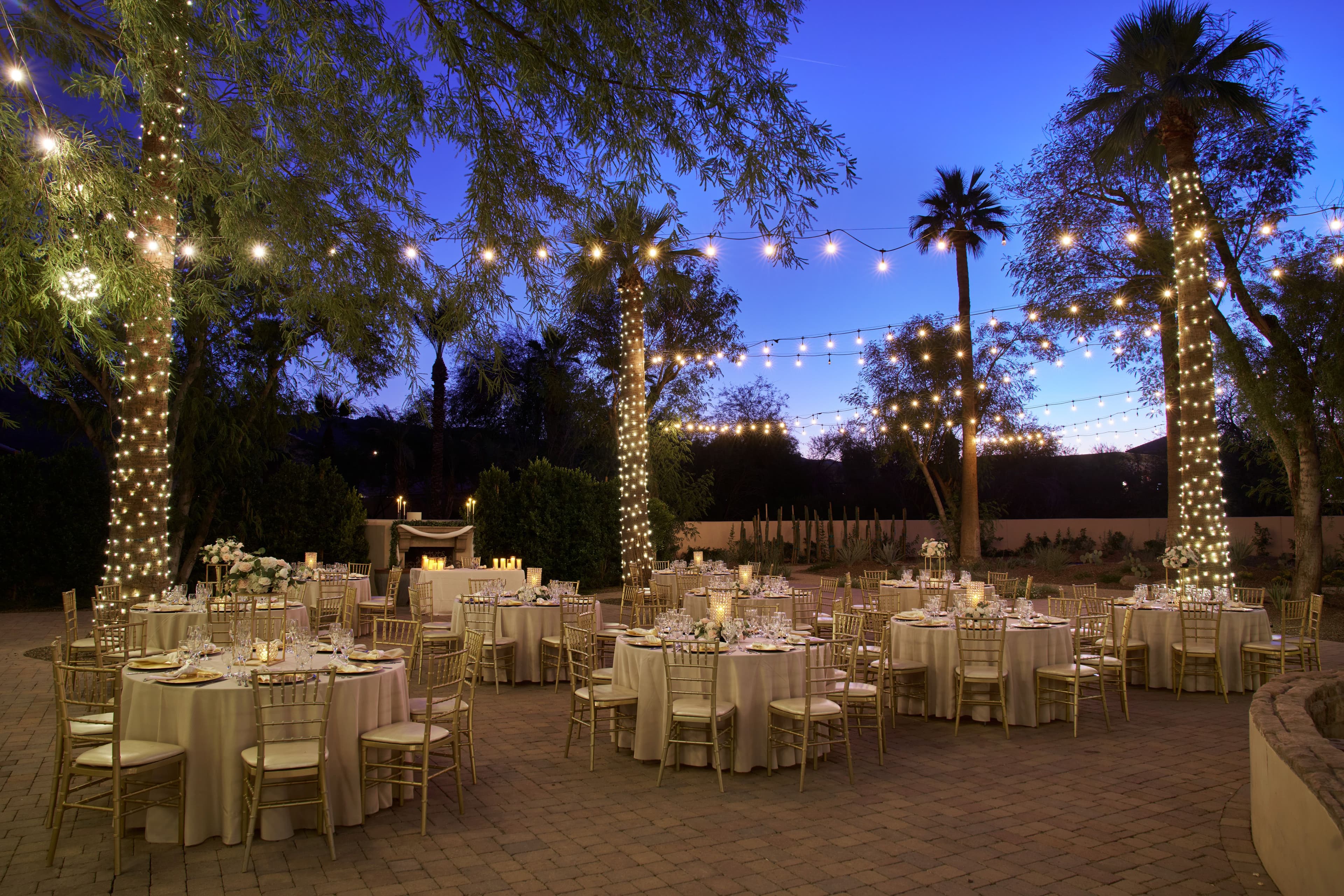 Romantic twilight wedding reception with palm trees wrapped in string lights and tables arranged on stone patio at Secret Garden
