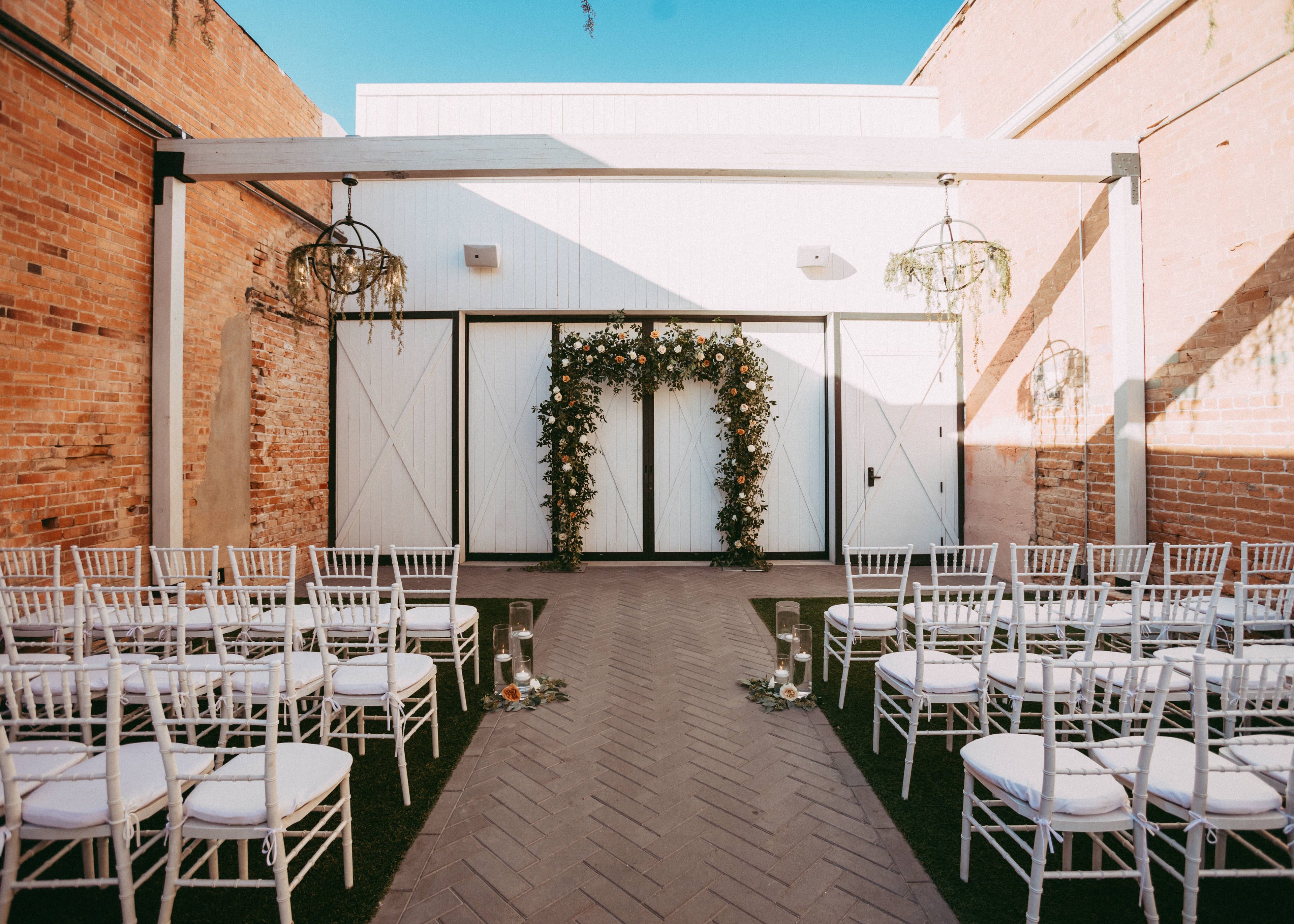 A rectangular floral arch frames the altar in this outdoor ceremony space. The white chairs fill the turf for guests. The space is surrounded by chic brick walls and wood beams.