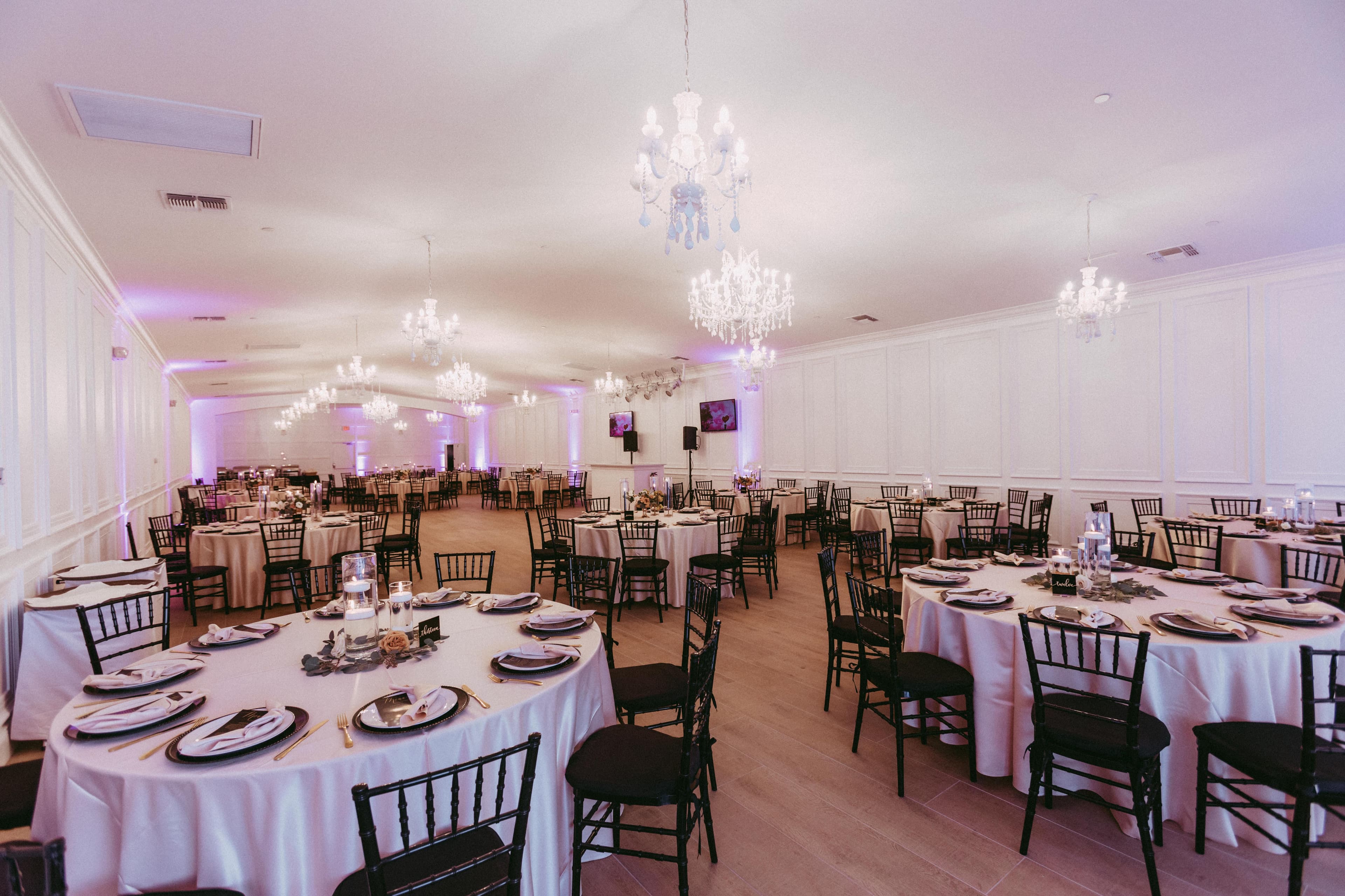 Alt text: Elegant wedding reception hall with chandeliers, round tables draped in white linens, black chairs, and soft purple uplighting creating a modern and romantic atmosphere.