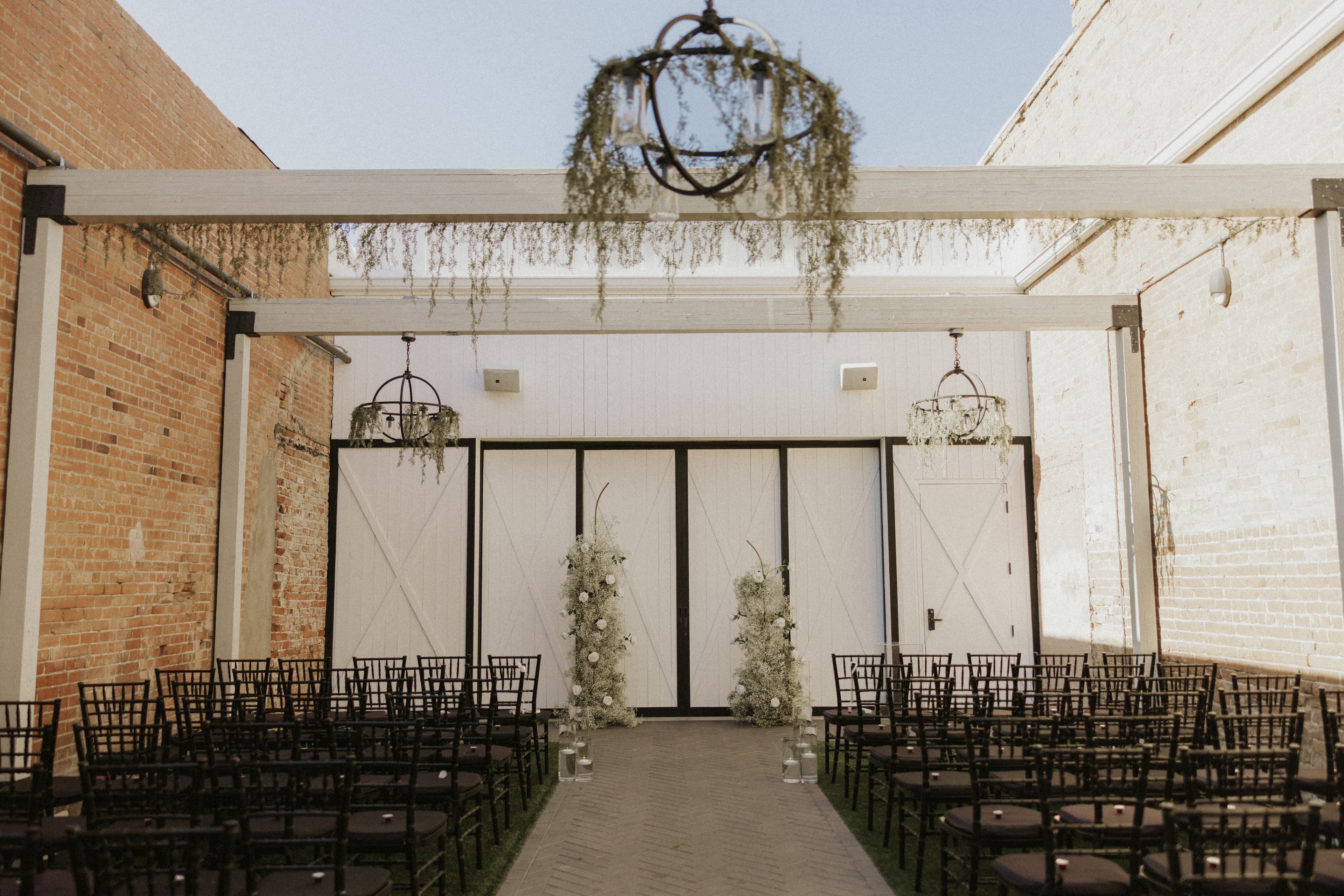 An open air space surrounded by brick walls and wood beams overhead. The space features a paver lined aisle and black charis all pointed at floral structures framing the altar.